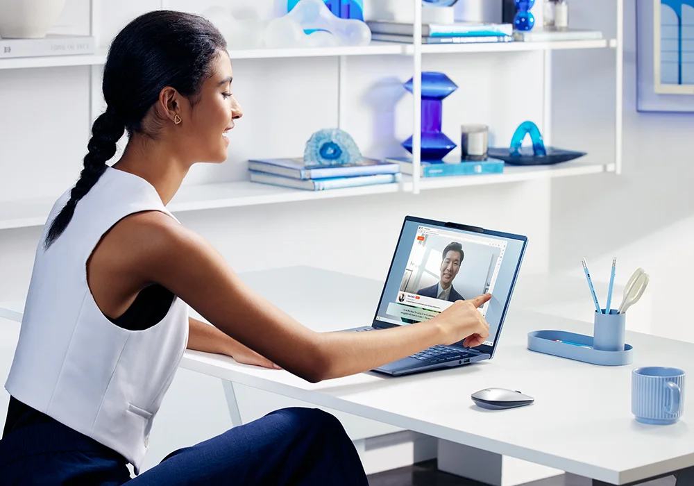 Person pointing at laptop screen during a video call, seated at a desk with office supplies, in front of shelves with blue decor and books.