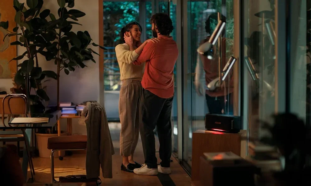 A couple smiling and embracing in a warmly lit, modern living room with large windows, indoor plants, and cosy furniture. The LG slogan 'Life's Good.' is prominently displayed, conveying comfort, connection, and contemporary lifestyle.