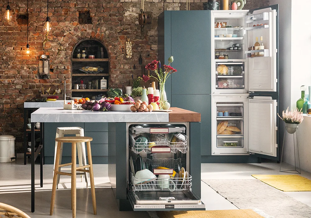 Modern kitchen with a person using a wall-mounted rangehood above an induction cooktop, which has a steaming pot on it; various kitchen items and plants are arranged on the countertop.