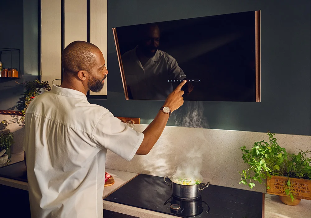 Modern kitchen with a person using a wall-mounted rangehood above an induction cooktop, which has a steaming pot on it; various kitchen items and plants are arranged on the countertop.