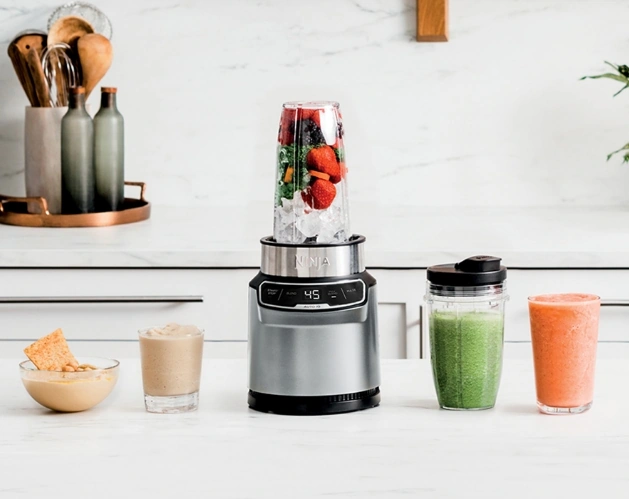 Ninja blender on a kitchen counter filled with ice, strawberries, and vegetables, surrounded by smoothies, dipping, and crackers. Showcasing versatile blending for healthy drinks and dips in a modern kitchen setting.