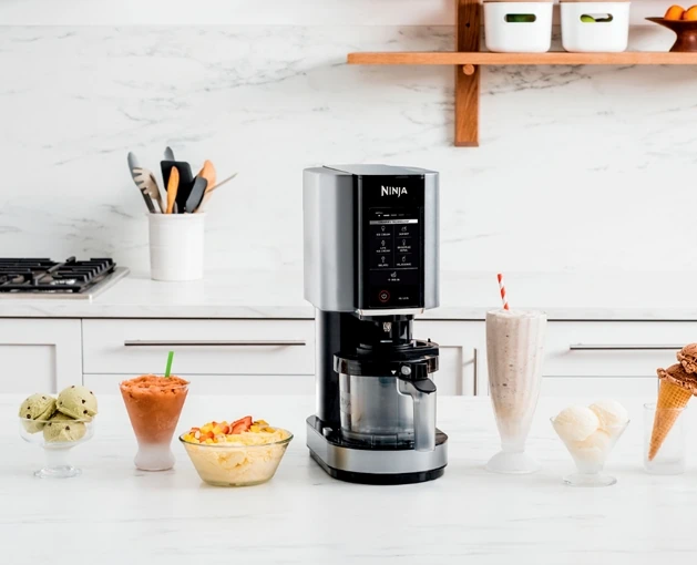 Ninja ice cream maker on a kitchen counter surrounded by frozen treats including ice cream scoops, milkshake, slushy drink, and fruit bowl. Showcasing a powerful and versatile dessert-making appliance.