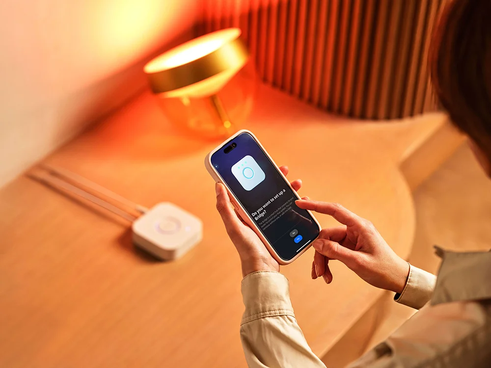 Person using a smartphone app to set up a Philips Hue smart device, with a warm-lit lamp in the background creating a cozy ambiance.