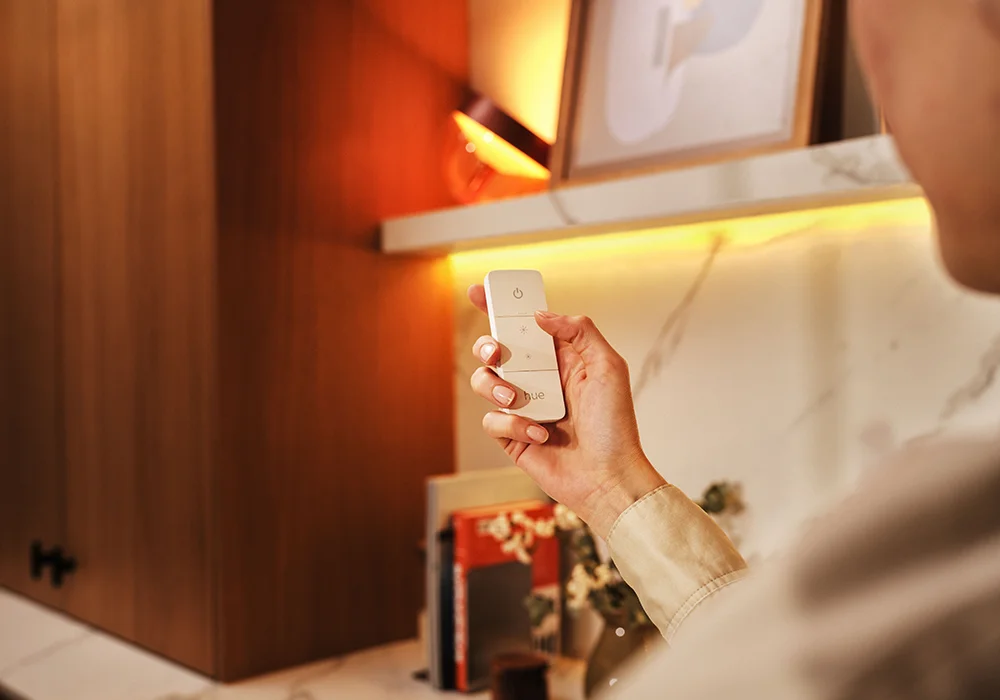 Hand holding a Philips Hue remote control in a warmly lit room with shelves, books, and wooden cabinetry, showcasing easy ambient light adjustment.