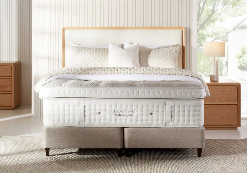 Crown Jewel Posturepedic mattress on a light grey textured mattress base, with plush pillows, wooden nightstands, and soft natural lighting.