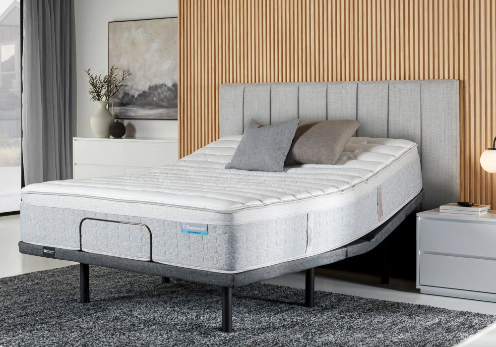 A Sealy Posturematic adjustable bed base with a grey upholstered headboard, layered pillows, and the Harrington mattress neatly made, positioned in a modern bedroom with wooden slat feature wall, bedside table with lamp, and a textured grey rug.