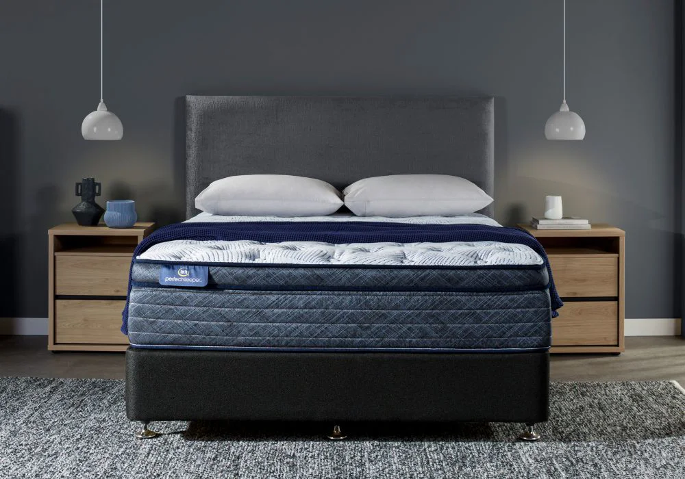 Bedroom with a Perfect Sleeper mattress dressed in white pillows and a dark blue blanket. Grey upholstered headboard, wooden nightstands, and pendant lights complete the modern setup.