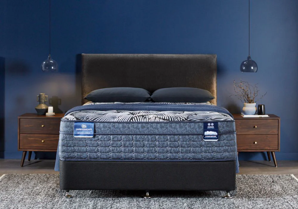 Perfect Sleeper X mattress with blue quilted design on a bed with a dark, velvet headboard, styled in a modern bedroom with dark blue walls.