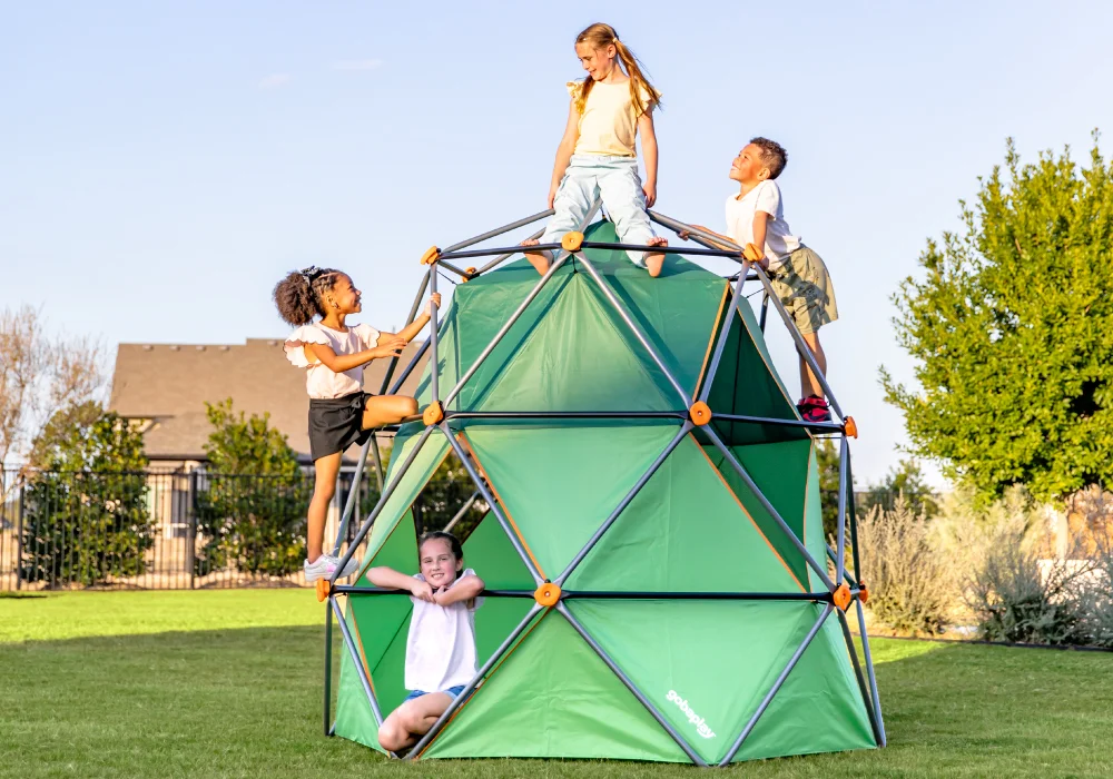 Four children playing on a green geometric climbing structure with orange connectors in a grassy outdoor area.