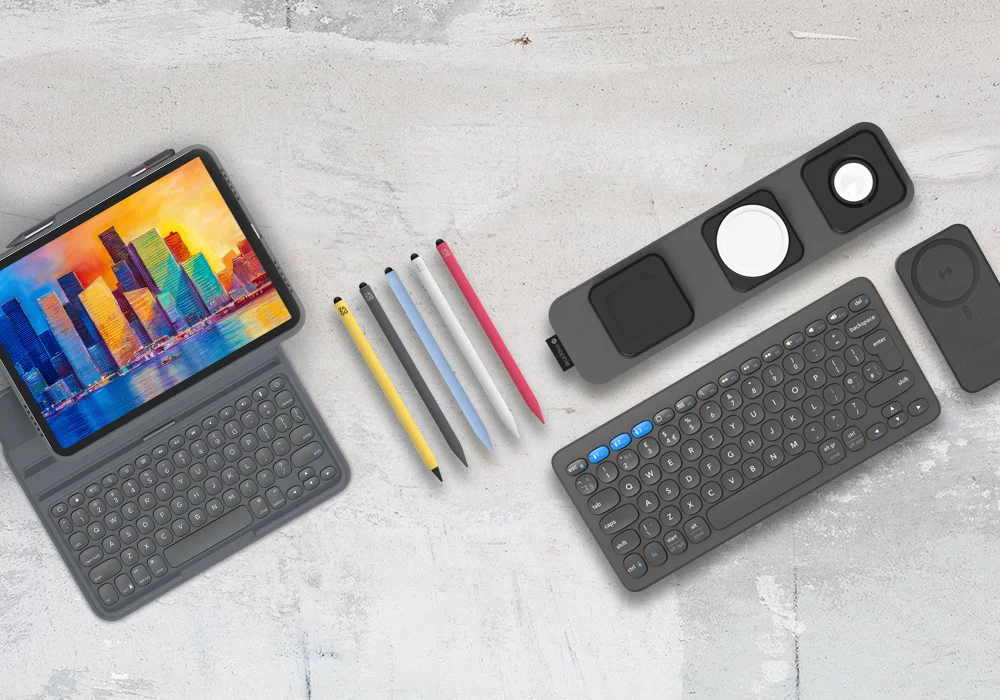 Tablet with a keyboard case, four colored pencils (yellow, blue, red, and white), a wireless charging pad with multiple slots, and a separate wireless keyboard. The tablet screen shows colorful buildings against a bright sky.