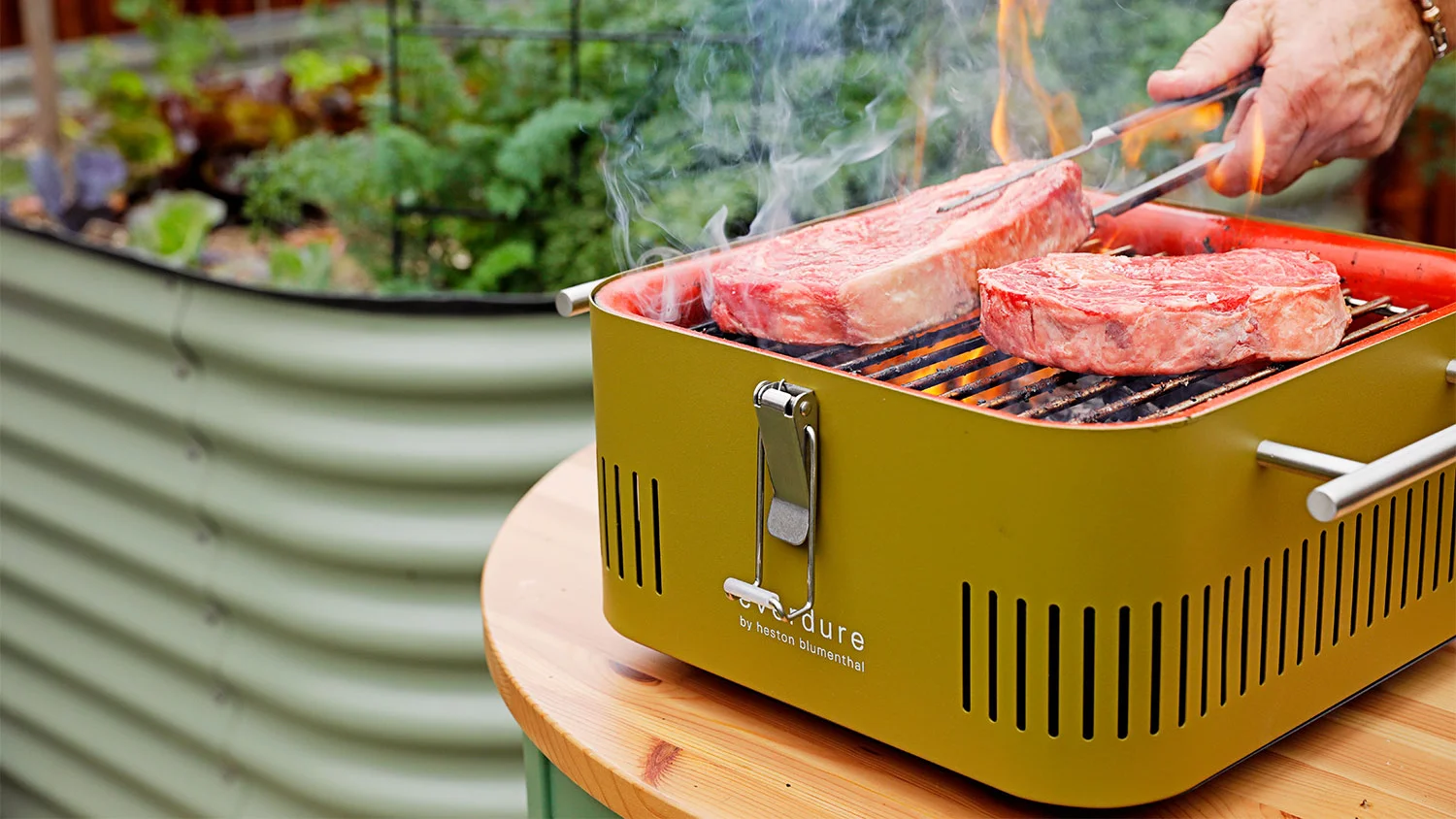 A person grilling steaks on the Everdure Cube portable charcoal BBQ by Heston Blumenthal, on a wooden table next to a raised herb garden.