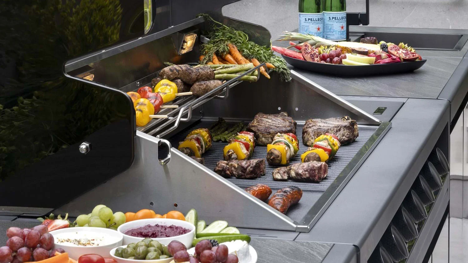 The Galaxy Gen2 BBQ warming racks loaded with grilled meats, skewers, and sides, surrounded by fresh produce and drinks for outdoor entertaining.