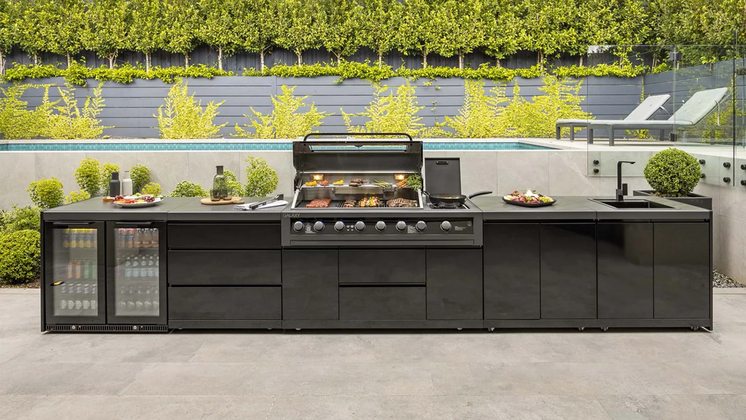 A Gasmate Galaxy Gen2 6-burner BBQ with built-in modules including a fridge, sink and prep stations in a modern outdoor kitchen beside a pool and lush garden.