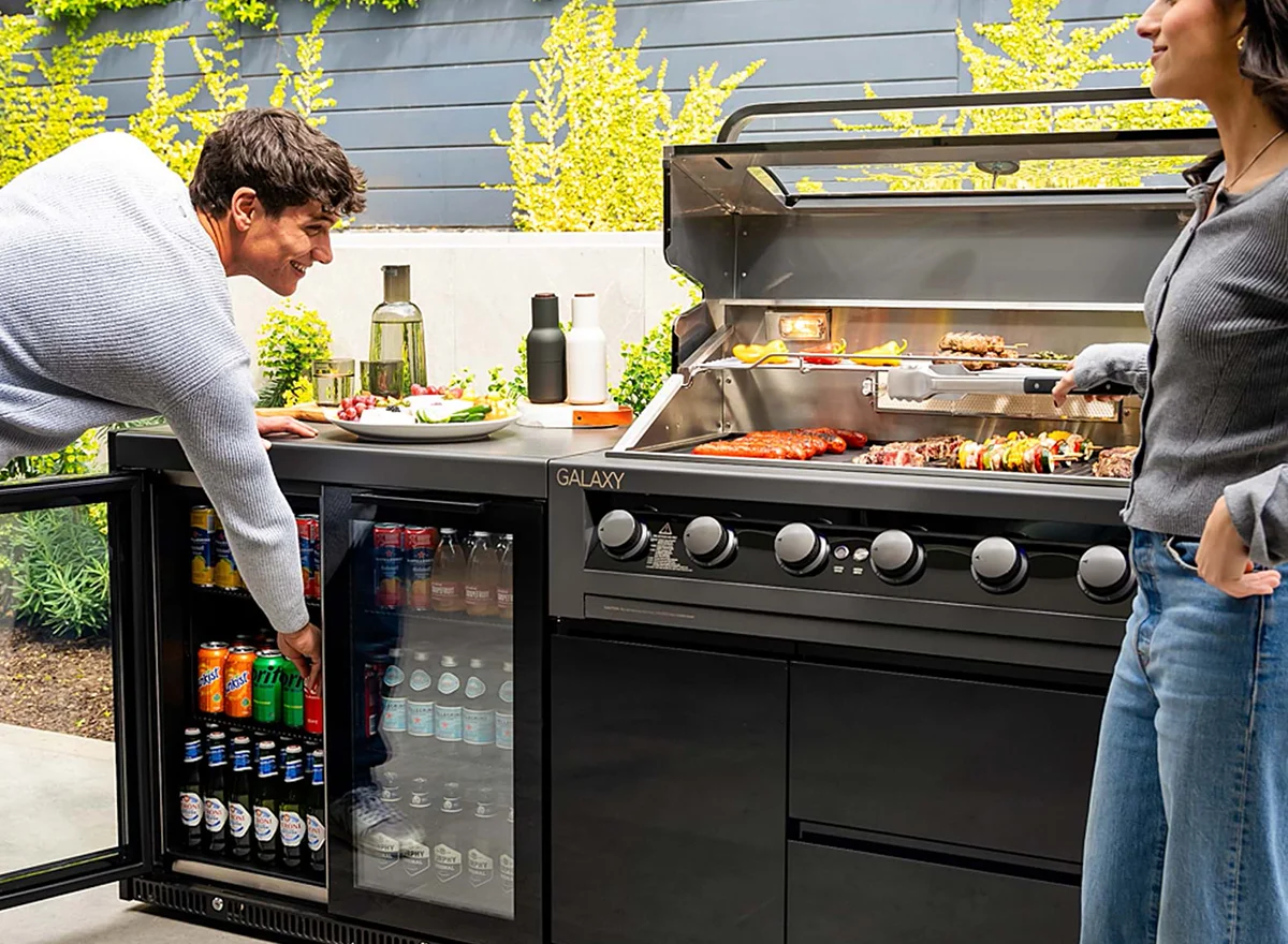 The Gasmate Galaxy Gen2 6-burner BBQ with built-in fridge in a modern outdoor kitchen, featuring a couple grilling food and grabbing drinks amid a garden backdrop.