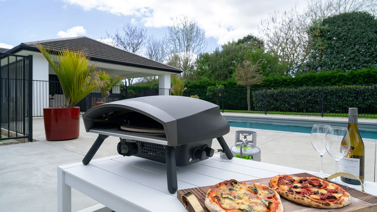 Outdoor pizza oven on a white table by a pool with two freshly baked pizzas, wine glasses, and a bottle of white wine in a modern backyard.