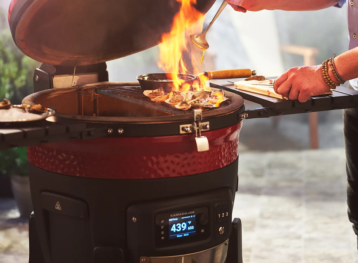 A Kamado Joe Konnected Joe ceramic BBQ with digital display showing 439°F, open lid, active flame, and grilling tools on side shelves during outdoor cooking.