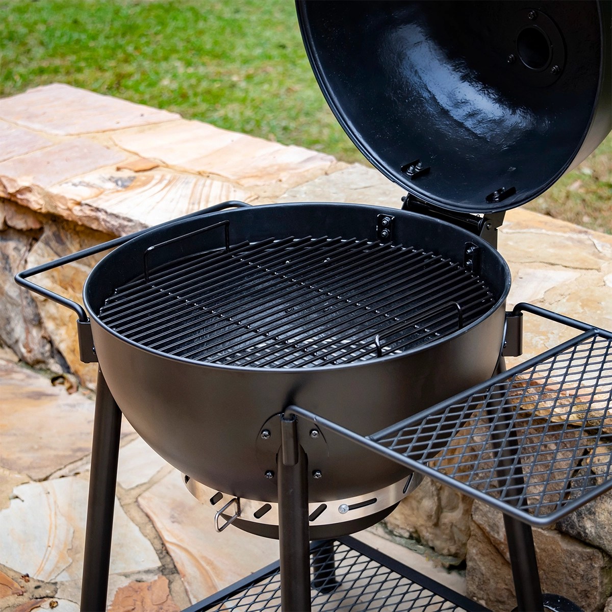 Black Oklahoma Joe's kettle charcoal BBQ grill with open lid, round cooking grate, and side mesh shelf, positioned on a stone patio with grass in the background.