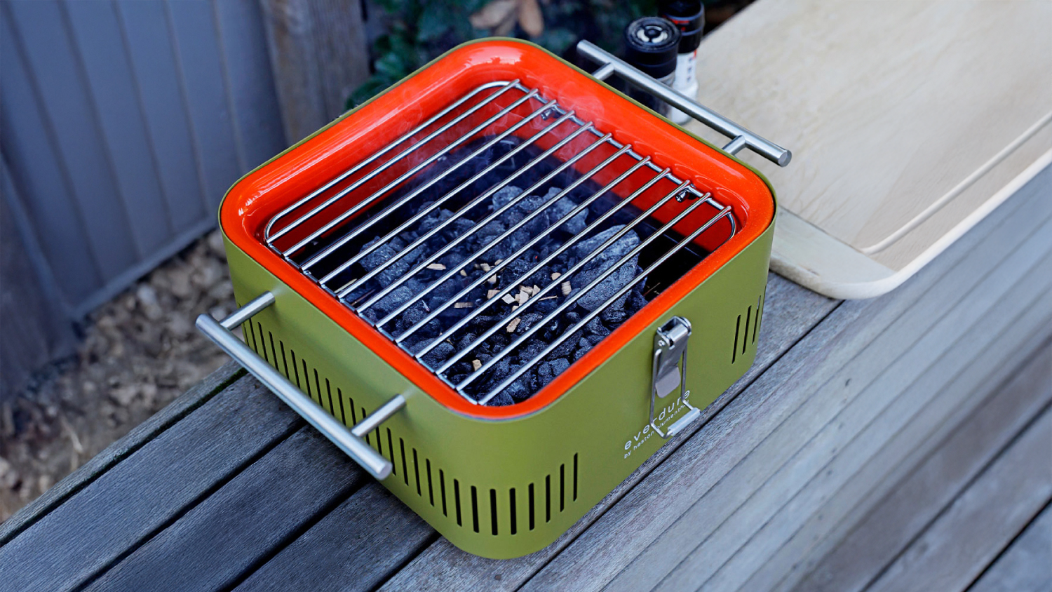 An Everdure Cube portable charcoal BBQ in green with lit coals and cooking grate, set on a wooden table beside condiments and a cutting board.