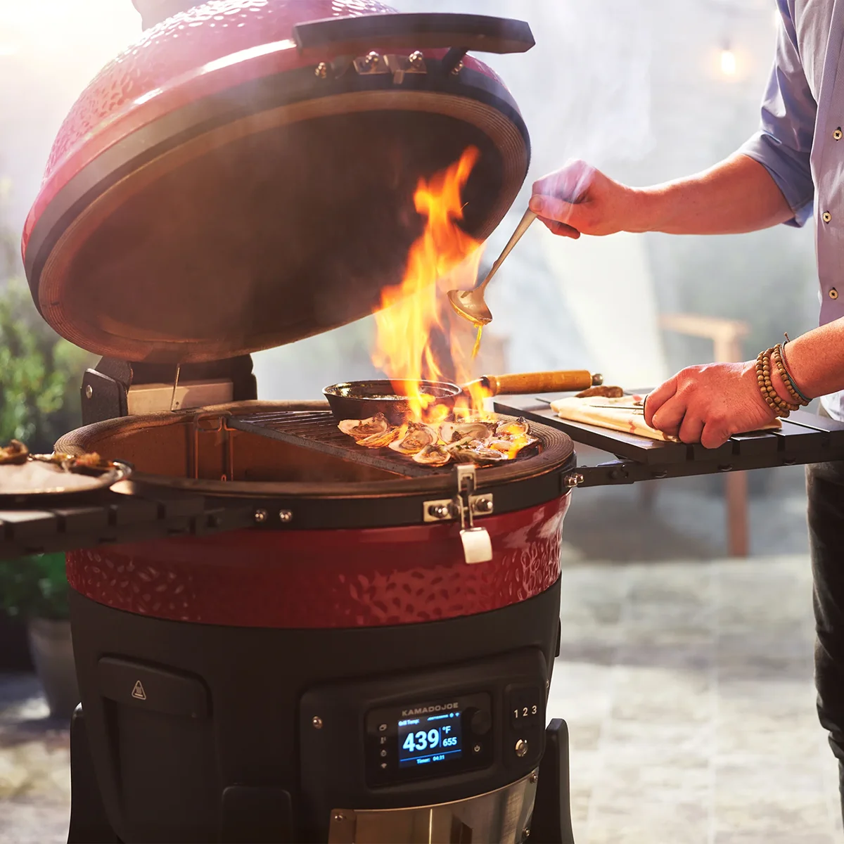A Kamado Joe Konnected Joe ceramic BBQ with digital display showing 439°F, open lid, active flame, and grilling tools on side shelves during outdoor cooking.