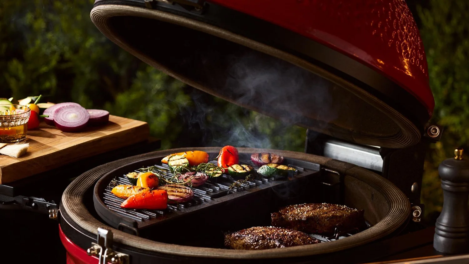 A Kamado Joe Classic II Red charcoal BBQ with the lid open, showing food cooking on the grill grates inside, set on a wooden deck with outdoor furniture and greenery in the background.