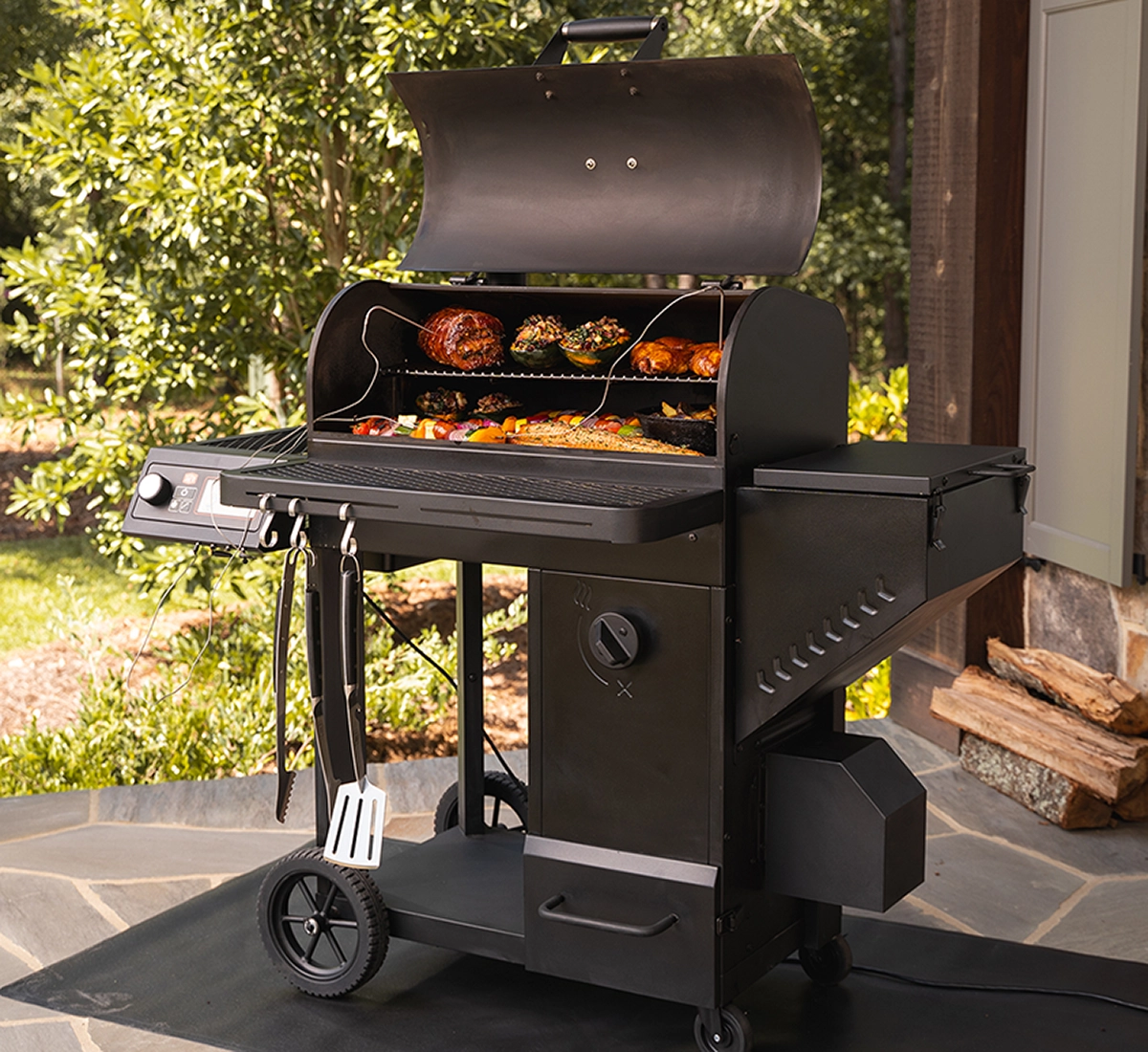 A Black Oklahoma Joe's Tahoma 900 auto-feed smoker grill on a patio, lid open showing assorted smoked meats and vegetables inside, with grilling tools hanging on the side, set against a green garden background.
