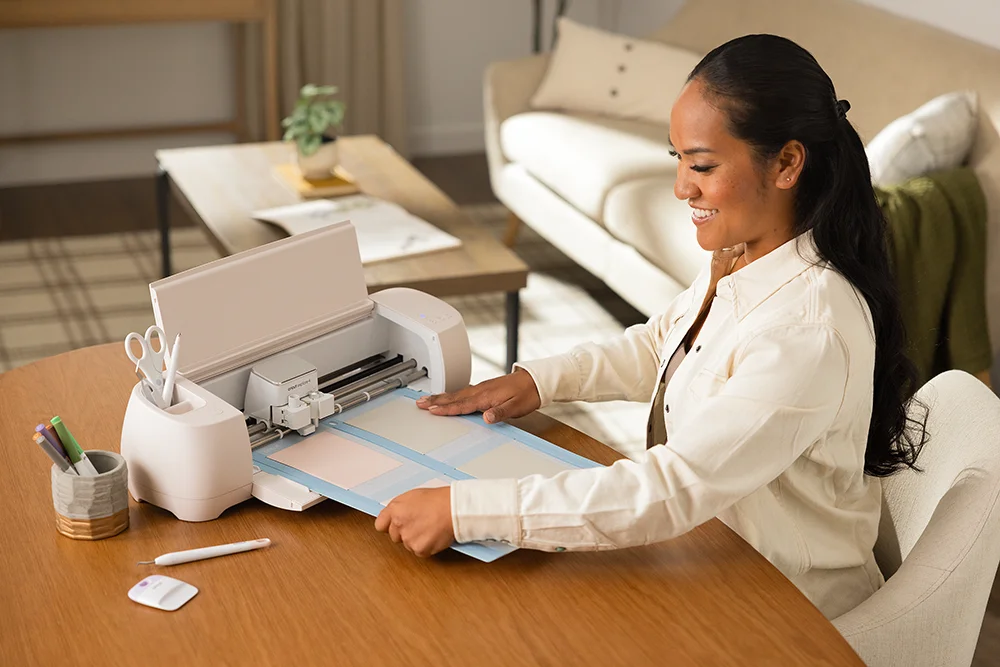 A woman sitting at a living room table running a print through a Cricut Explore 4 machine.
