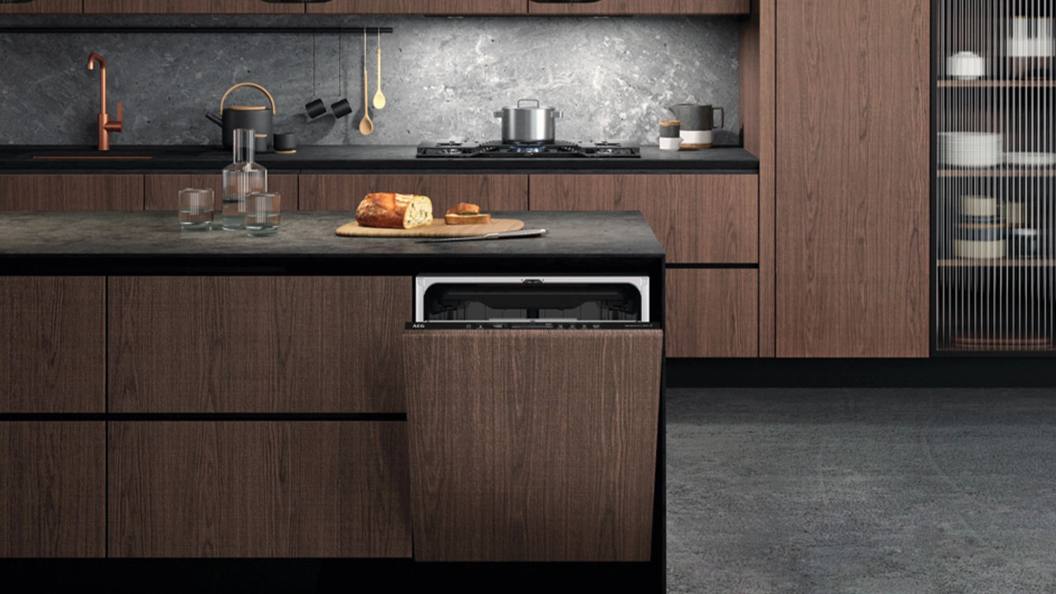 An AEG fully integrated dishwasher with wood-panel front partially open in a modern kitchen featuring dark wood cabinetry, a stone benchtop, a gas cooktop with cookware, and a cutting board with bread and glasses arranged on the island.