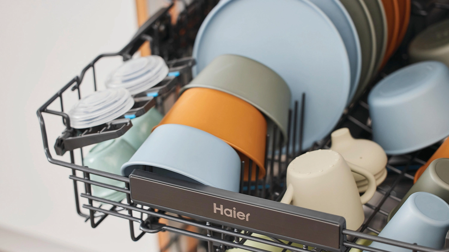 The upper rack of a Haier dishwasher loaded with assorted dishes, including pastel-colored bowls, cups, and plates, including a sippy cup securely arranged in dark grey tines with a dedicated holder for small lids on the left side.