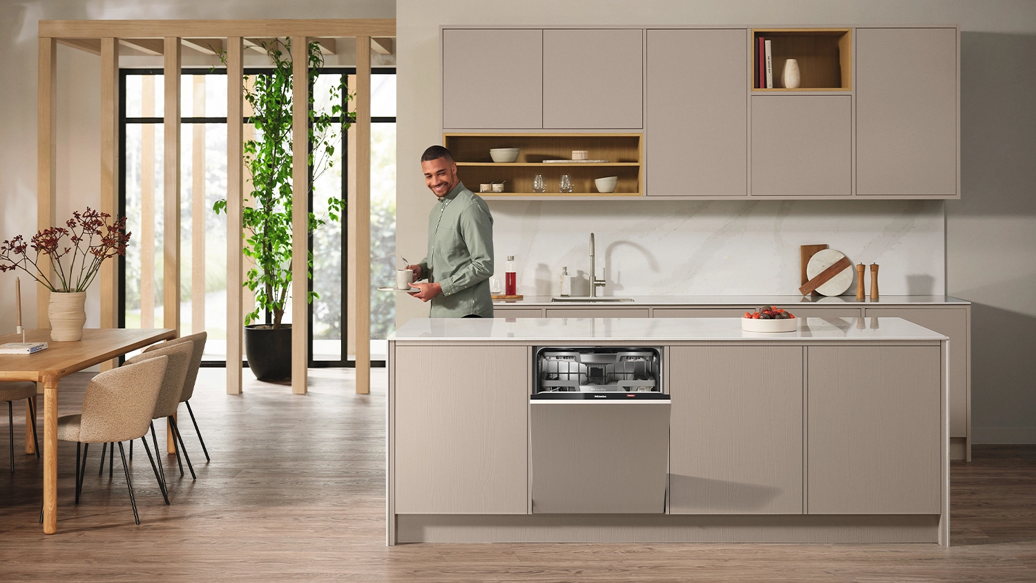 Modern kitchen with light cabinetry and a central island featuring a Miele 15 place setting  fully integrated dishwasher with the door partially open, under a white laminate island with a fruit bowl on top. In the back is a marble-look splashback, open shelving with dishes above the sink, and a dining area on the left with wood furniture and large floor-to-ceiling windows.