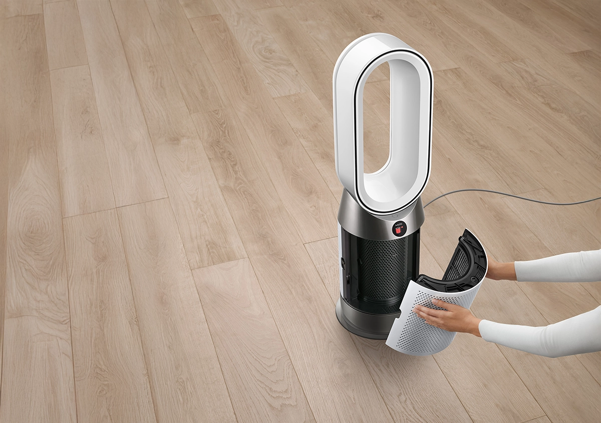 Dyson HP11 Purifier Hot+Cool fan heater in silver and white placed on a wooden floor, with a person's hands installing or removing the air filter from the base of the unit.