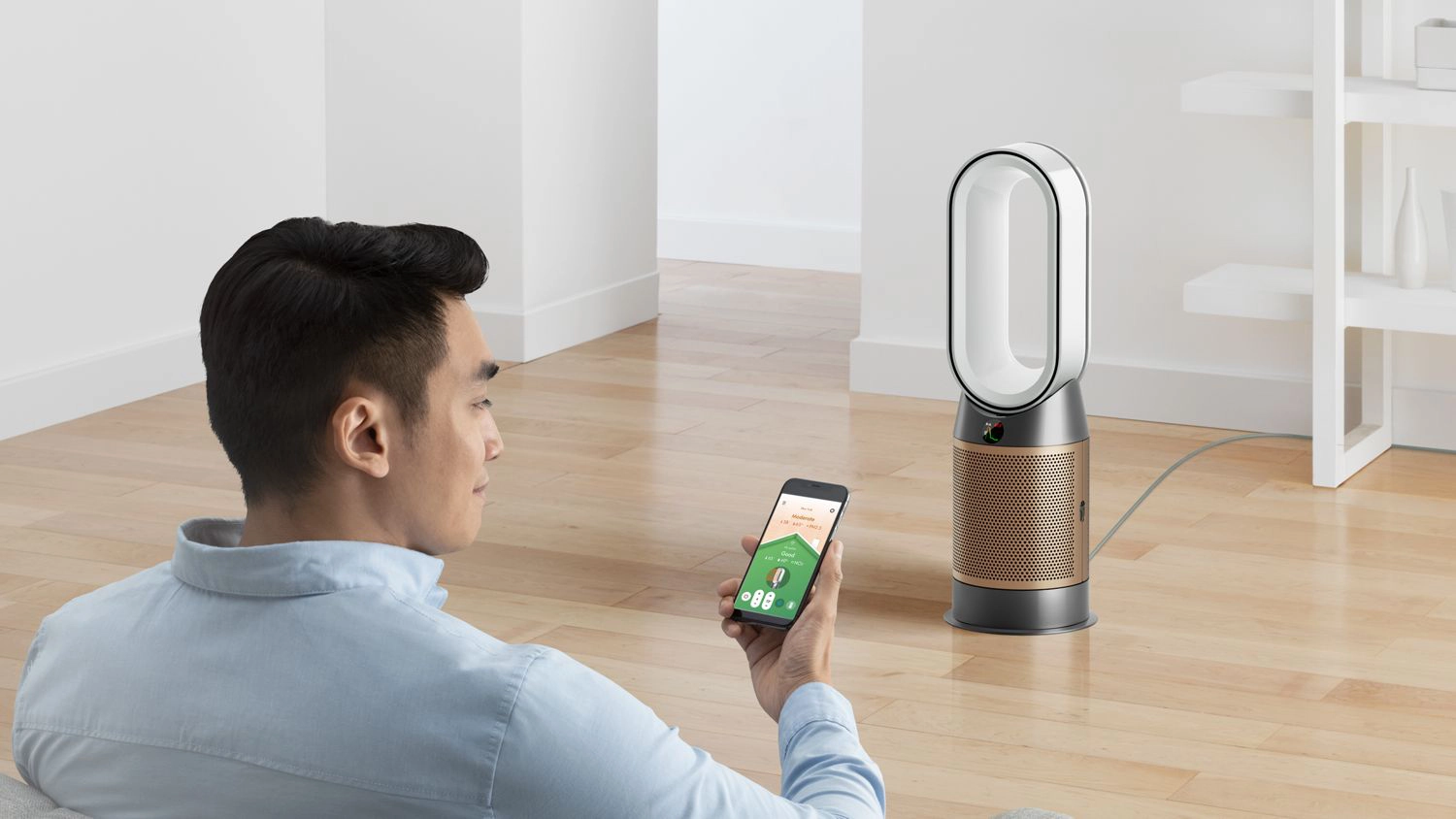 A Dyson HP12 Purifier Hot+Cool fan heater with sleek oval design and gold-toned base placed on a wooden floor, shown next to a person holding a smartphone displaying the Dyson app in a modern living room