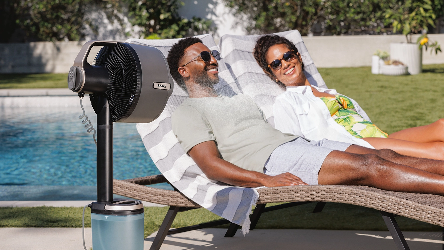 A Shark FlexBreeze Pro Mist indoor-outdoor pedestal fan cooling  two people relaxing on wicker lounge chairs near a swimming pool, with green lawn and garden in the background.