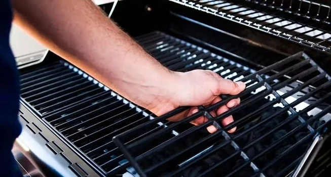 Close-up of a clean black BBQ grill with a person lifting the main grate, revealing a multi-level cooking surface including an upper warming rack.
