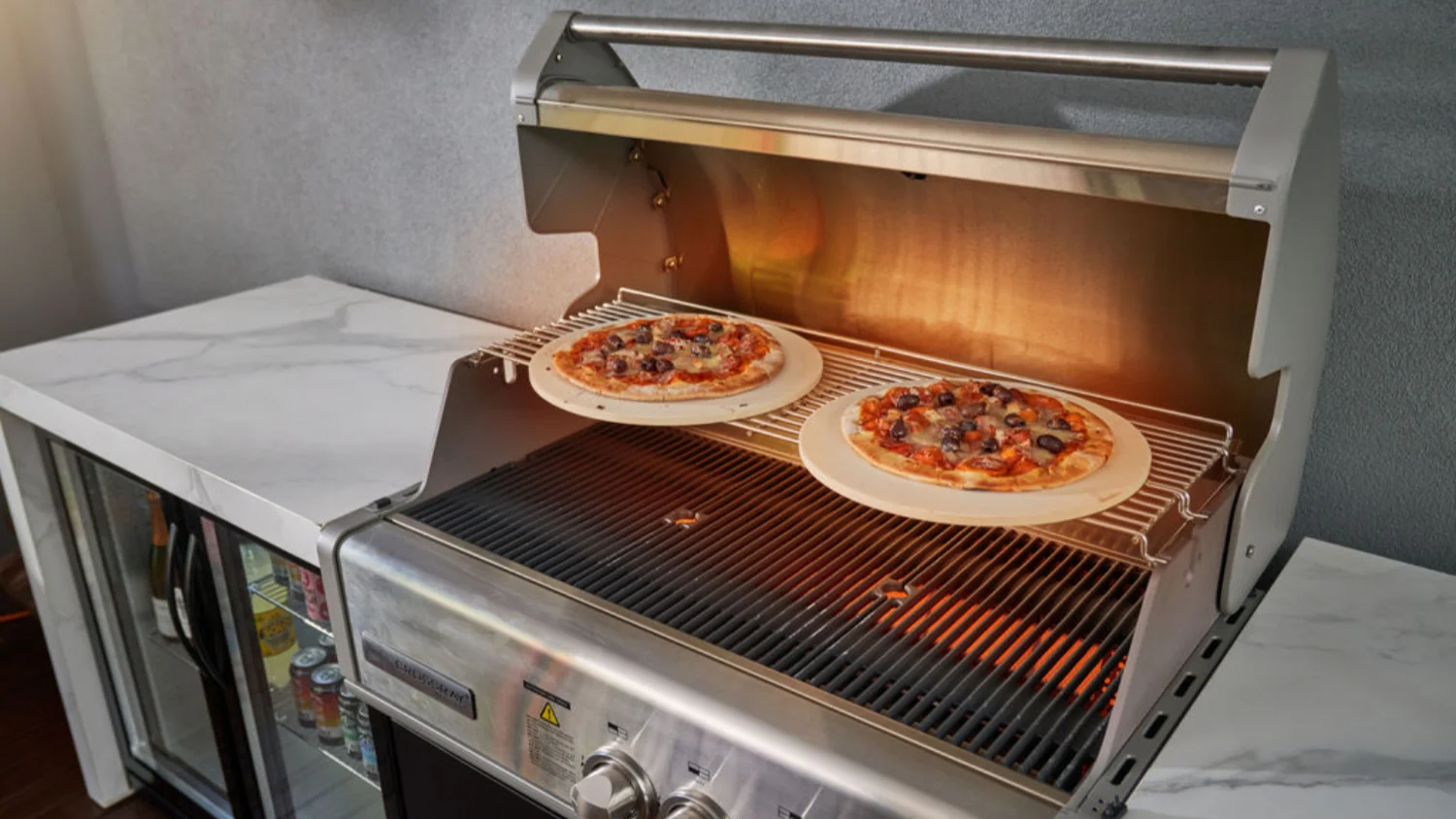 The Crossray gas BBQ featuring warming racks with a couple of pizzas cooking, a flat griddle and traditional grill grates with burners on.