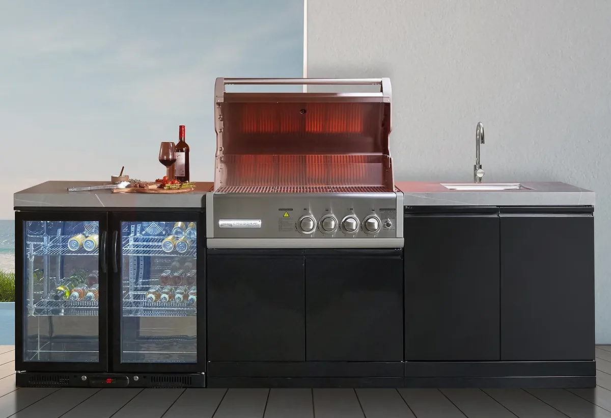 A Crossray 4B-S Series stainless steel outdoor BBQ kitchen with open grill, double-door beverage fridge, with food and wine on top, and built-in sink module, set against a coastal backdrop.