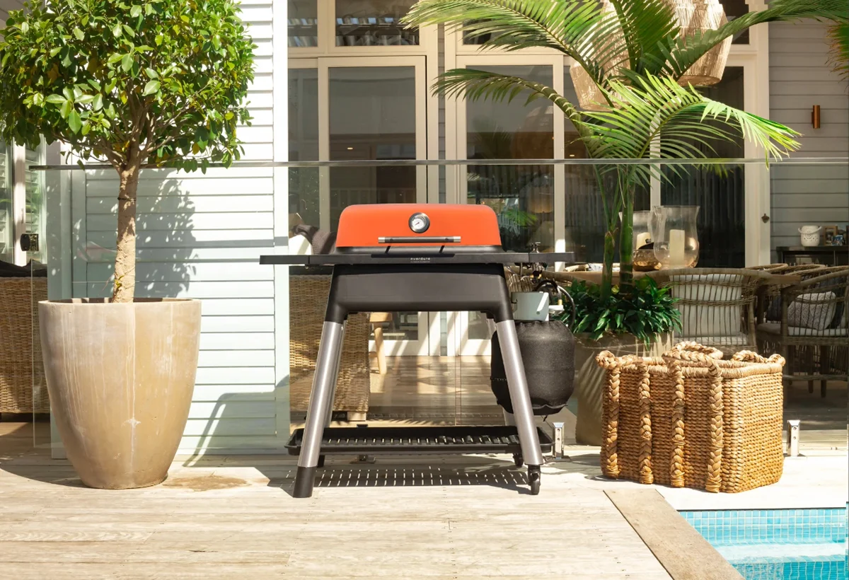 An Everdure 2nd Gen freestanding gas BBQ with orange lid on a black and silver stand, set on a sunlit patio surrounded by plants and a wicker basket next to a pool.