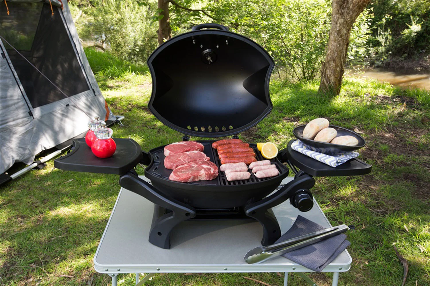 A Gasmate Odyssey portable BBQ on a camping table at a campsite with grilled meats, condiments, and buns on the side shelves, and a tent in the background.