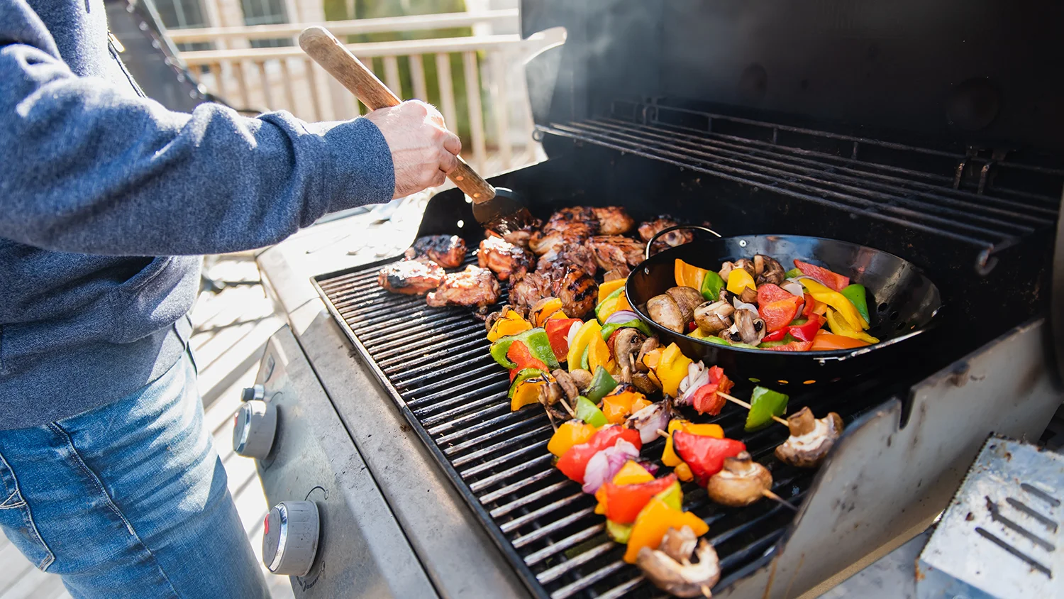 A Close-up of skewers with grilled meat, bell peppers, and onions cooking over an open flame on a barbecue grill, with a hand using tongs to turn the food in an outdoor setting, set on a stone patio.
