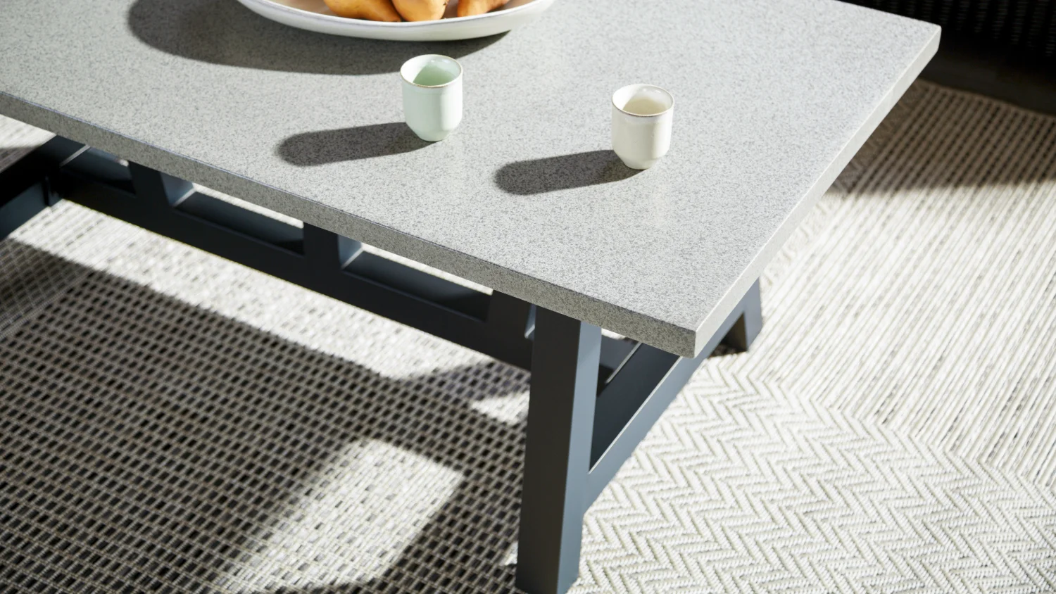 The light grey textured, concrete fibre board top and powder-coated aluminium legs of the Toulouse coffee table, on a woven herringbone rug in natural light.