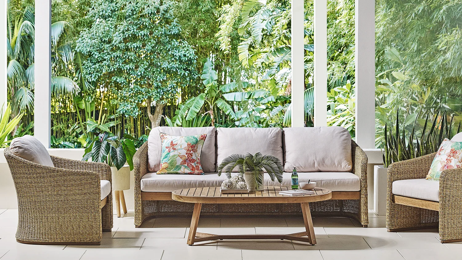 The Florida 7-piece outdoor lounge suite with PE wicker sofa and chair featuring light-colored cushions, a floral-patterned accent cushion, and a wooden coffee table holding a potted plant and green glass bottle, on the patio surrounded by lush green foliage.