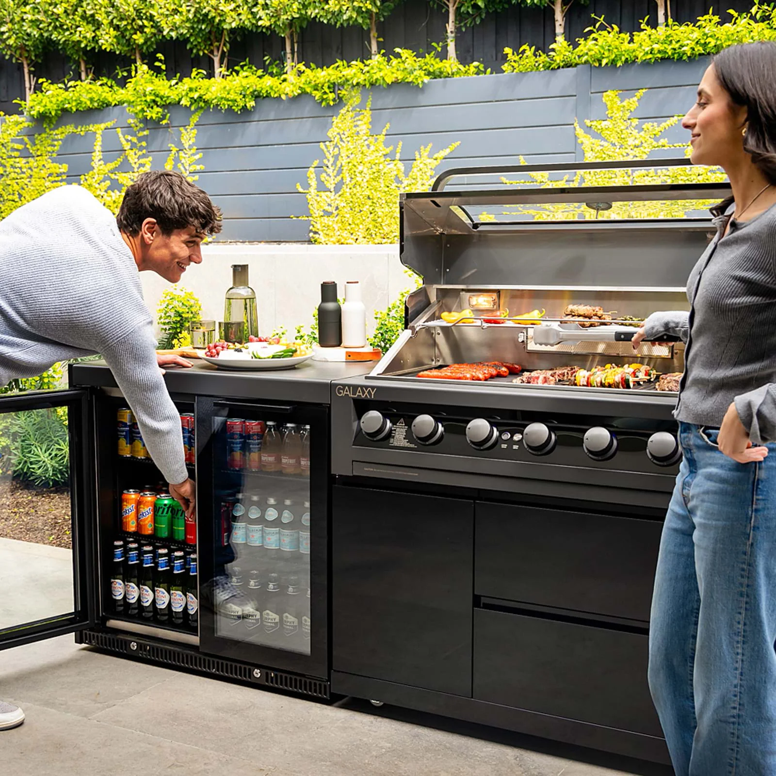 The Gasmate Galaxy Gen2 6-burner BBQ with built-in fridge in a modern outdoor kitchen, featuring a couple grilling food and grabbing drinks amid a garden backdrop.