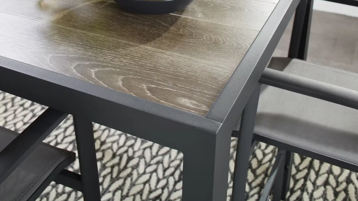 The wood-top table and dark metal frame chairs of the Wardell 5-piece outdoor bar setting on a black-and-white leaf-patterned rug.