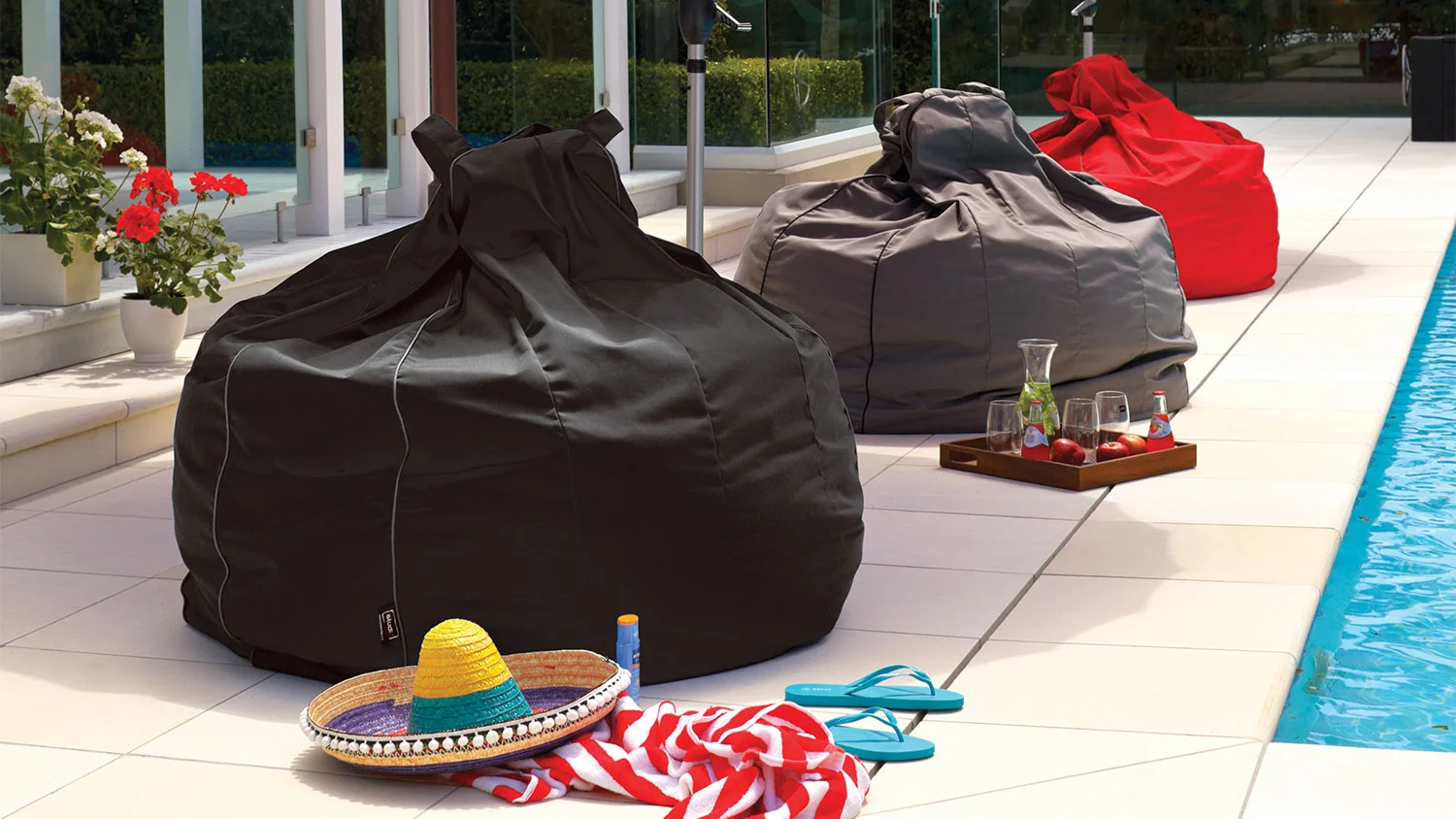 The Studio Jumbo Bean Bags in black, grey, and red by a poolside patio, styled with summer accessories, drinks, and potted flowers for a relaxed outdoor vibe.