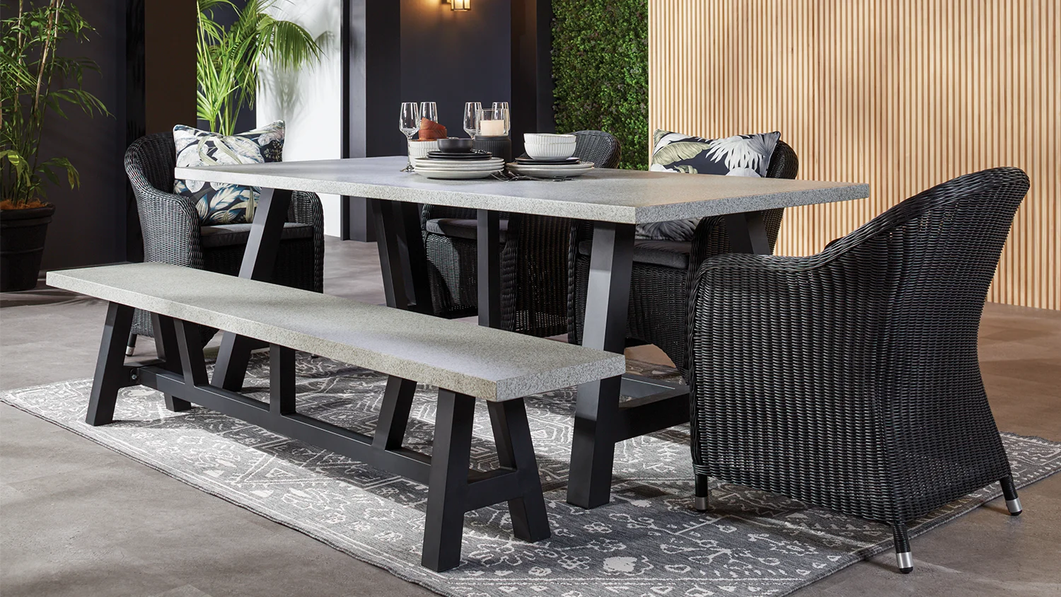 Toulouse 6-piece grey outdoor dining suite with cushioned wicker chairs, bench seating, and a rectangular table styled with tableware and plants on a patio with light siding and scenic views.