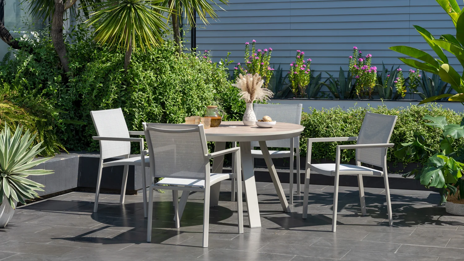 The Urbanite 5-piece outdoor dining suite in Coral Sand with round wooden table and chairs, styled with vases and bowls on a tiled patio surrounded by lush greenery.