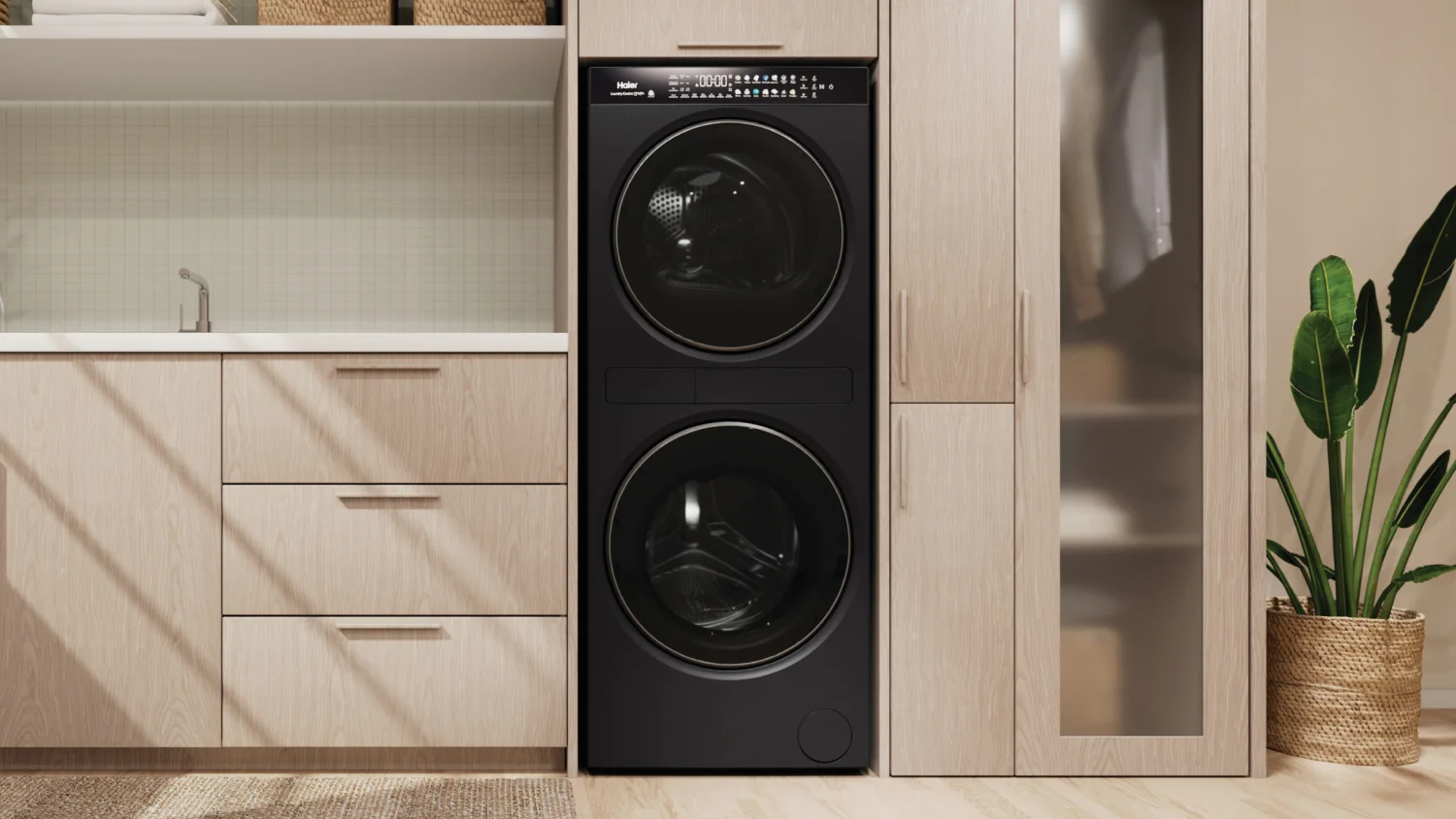 Modern laundry room with a black stacked washer and dryer built into light wood cabinetry, alongside a laundry sink, storage drawers, and a potted plant.