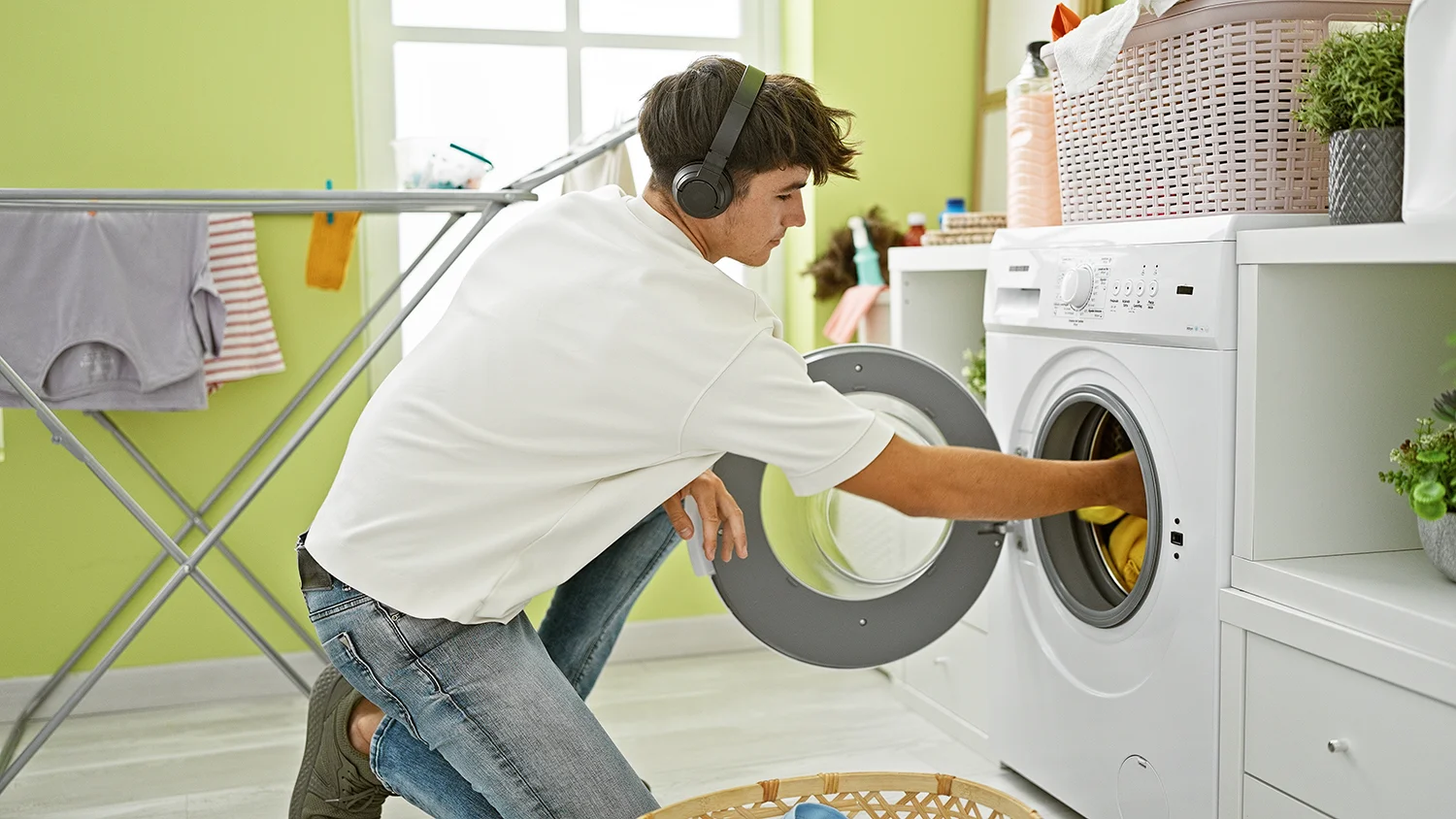 A teenage boy with headphones and casual clothing kneeling in front of a washing machine, loading or unloading laundry. A laundry basket, ironing board with hanging clothes, and shelves stocked with detergent and folded laundry suggest a well-organised space.