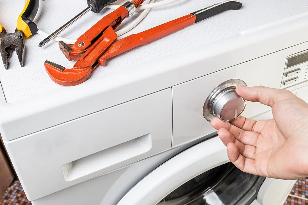 Close-up of a washing machine with tools on top and a hand turning the control knob, showing setup or repair during installation.