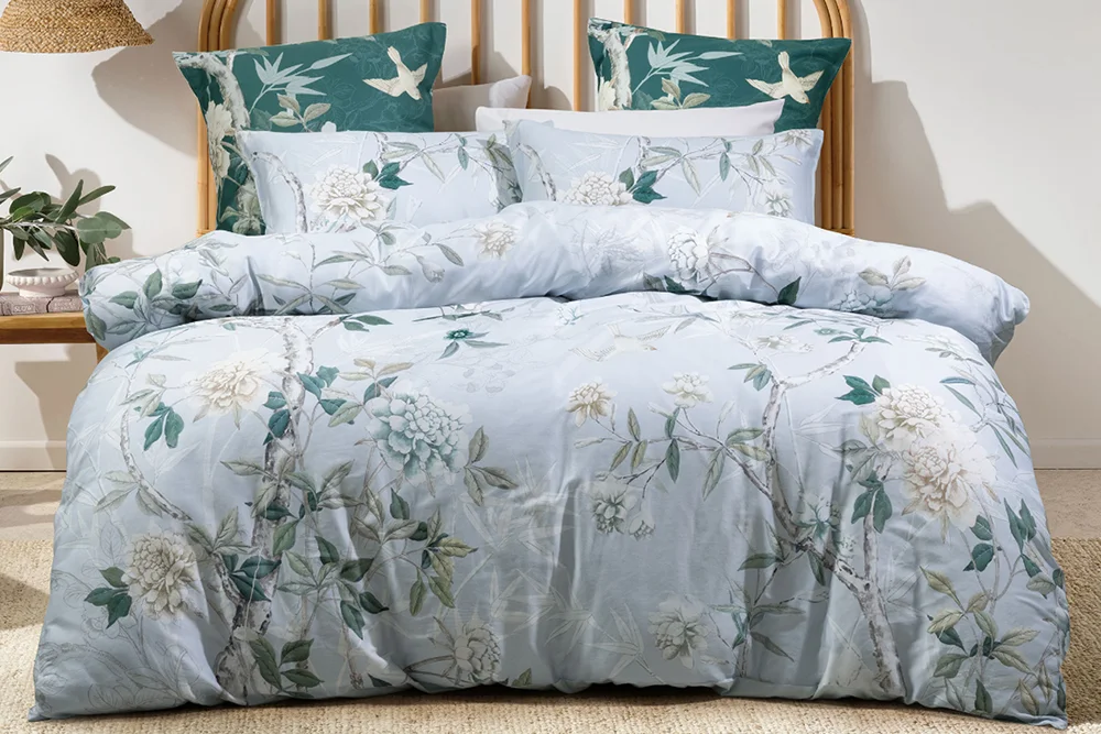 Bedroom with the Anika Duvet Cover Set by Savona, styled in light blue floral patterns with matching European pillows, a wicker headboard, and a bedside table featuring a pot plant and a hanging shade.