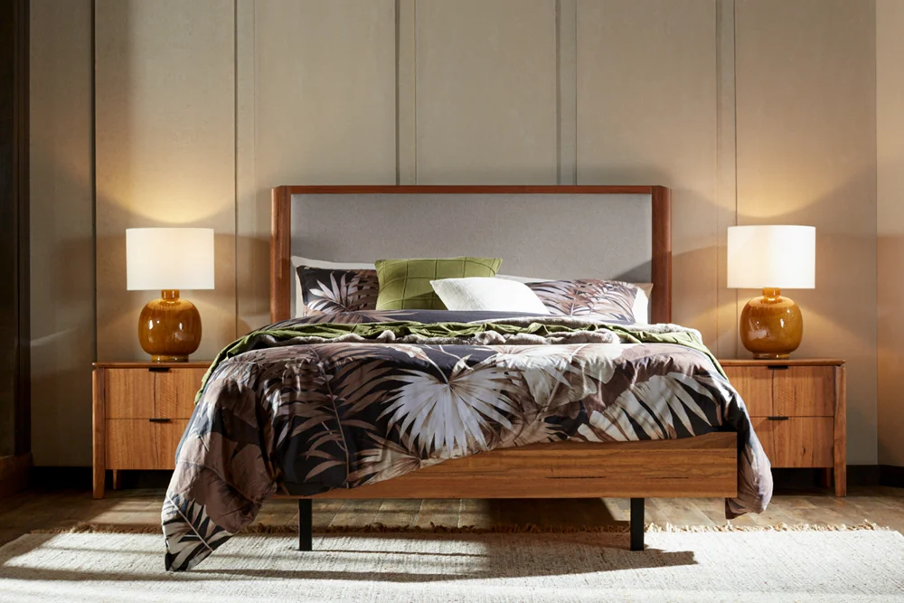 Tropical-style bedroom featuring the Cory Queen Upholstered Bed Frame, styled with palm leaf bedding, green and white pillows, with matching Cory bedside tables, and ceramic lamps with white shades.
